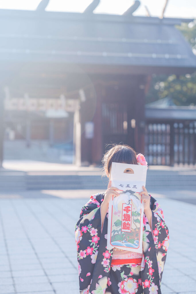 札幌護国神社を背景に千歳飴の袋で顔を隠して遊ぶ着物姿の七五三の女の子